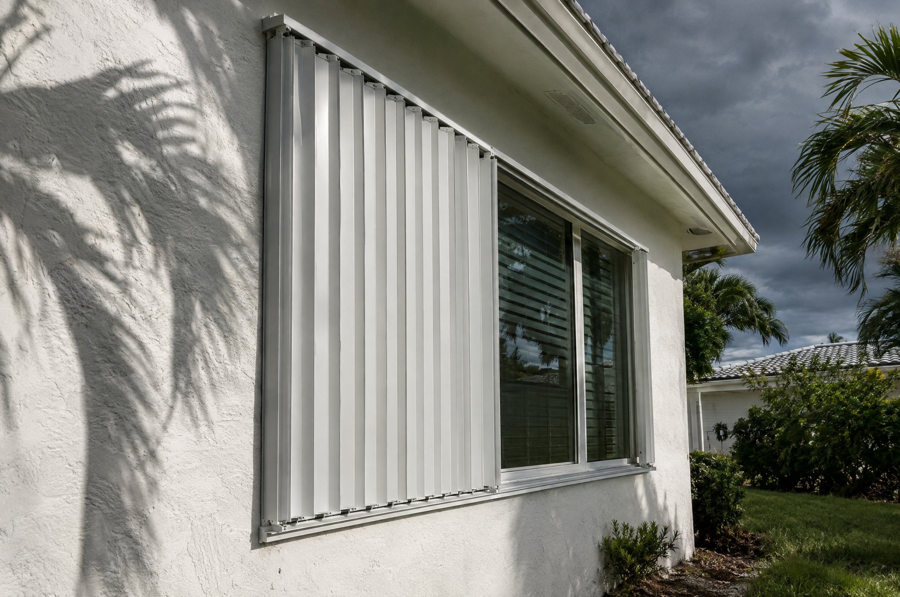 White aluminum storm shutter panels closed over a window on a white stucco Florida home under a gray sky