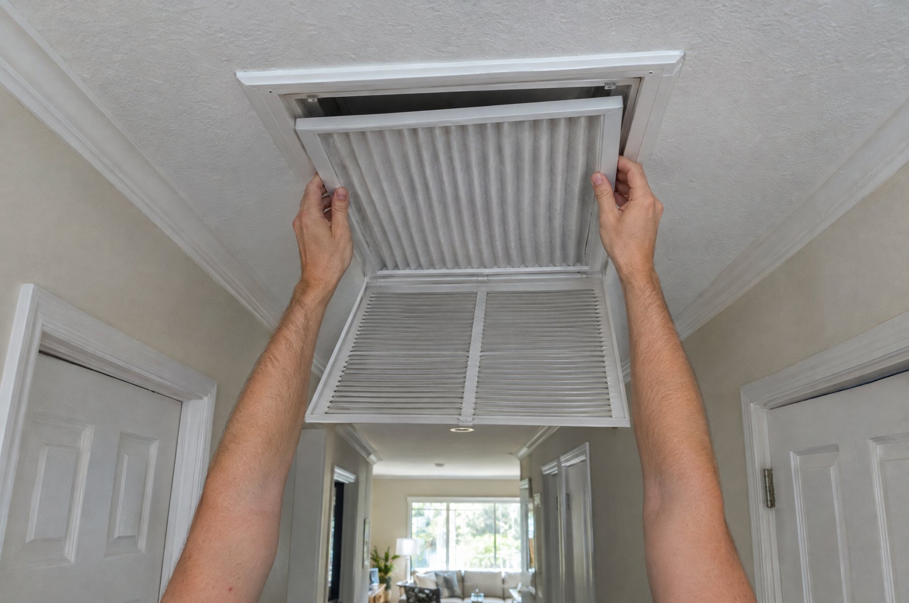 Hands pushing a new full-size white pleated air filter into a central return vent in a hallway ceiling, the hinged grille hanging open below