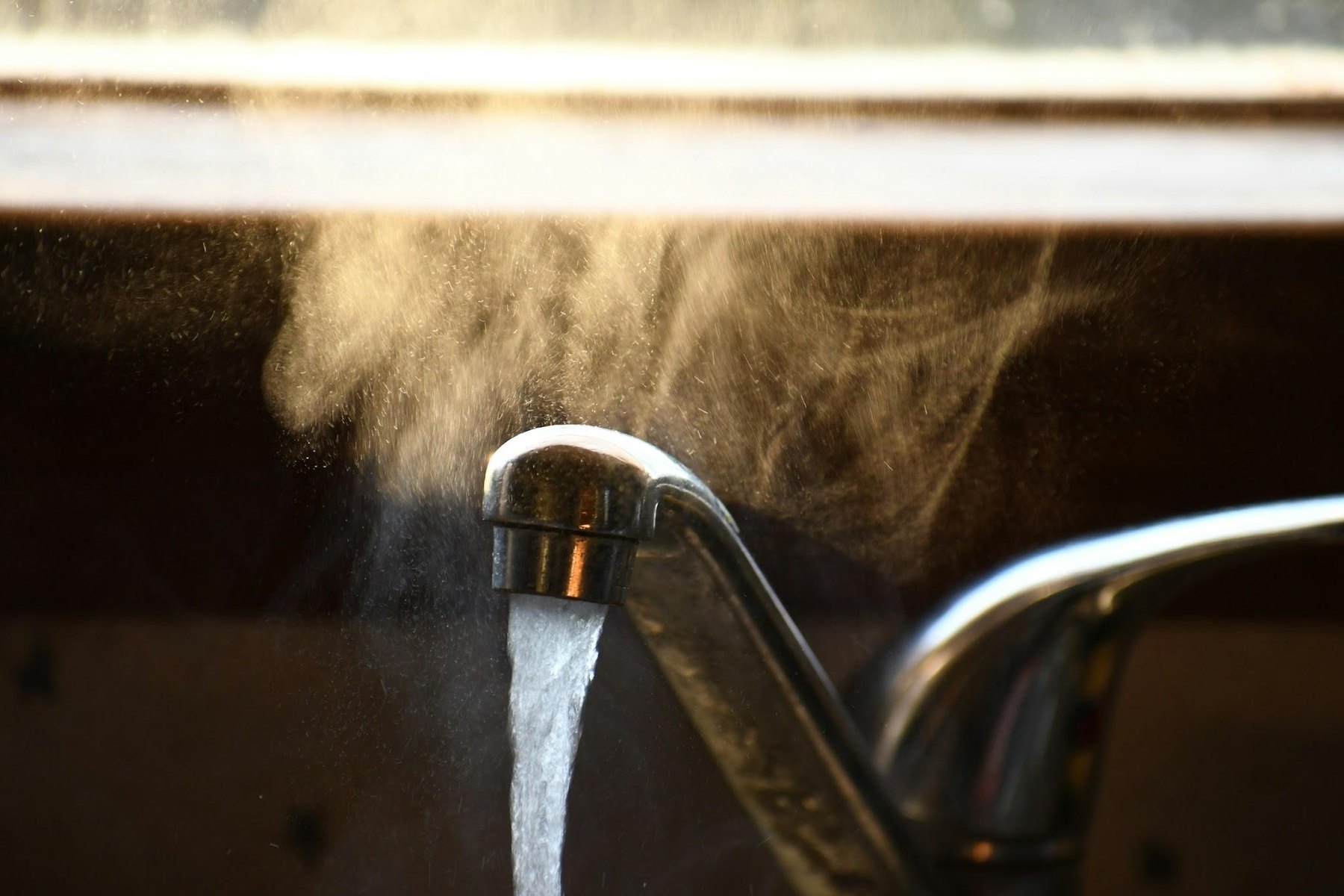 Steam rising from hot water running out of a chrome kitchen faucet in warm light