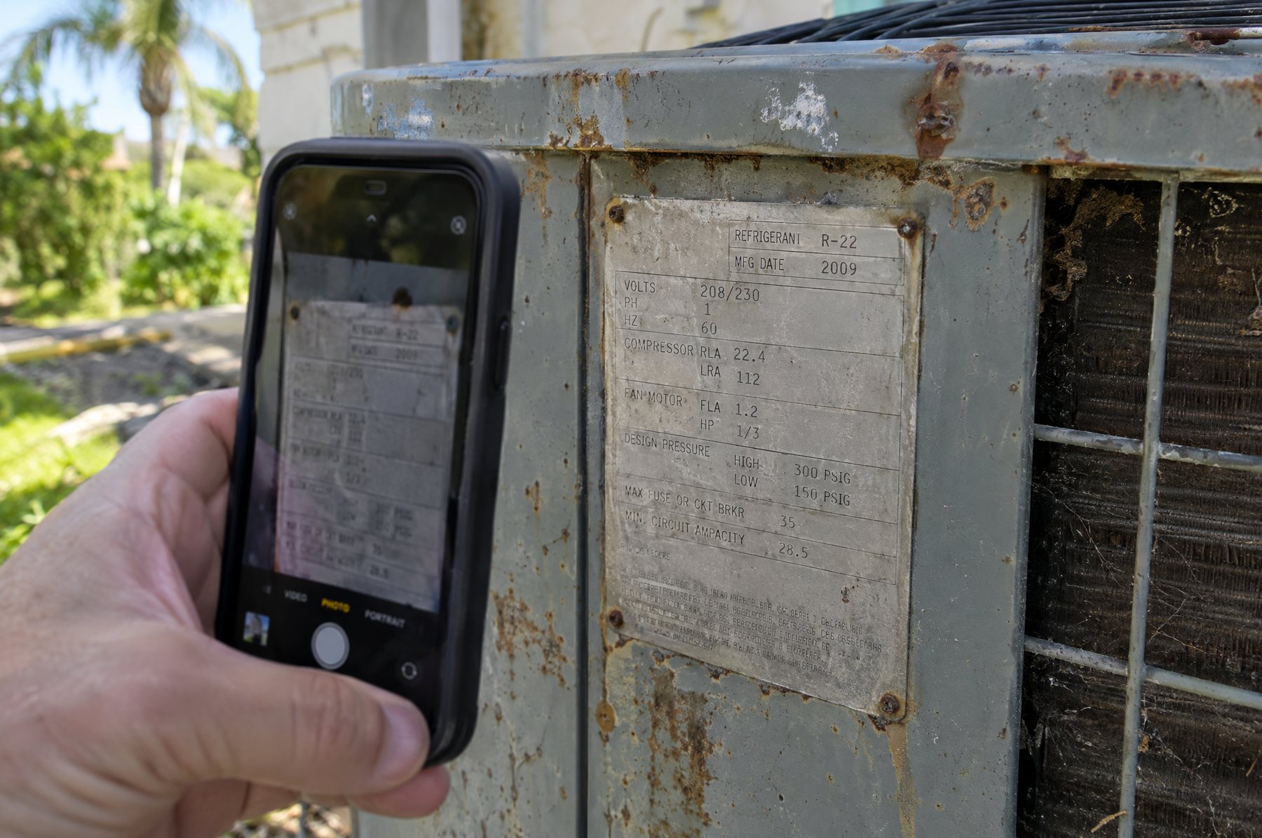A homeowner photographing the data plate on a weathered outdoor air conditioner, which lists the refrigerant as R-22 and a 2009 manufacture date