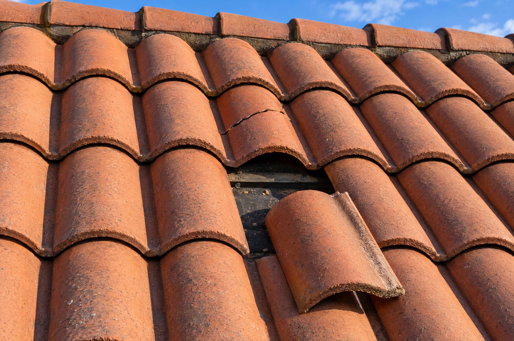 Close-up of a terracotta barrel tile roof with one tile cracked across the middle and another slipped out of its row, exposing the underlayment