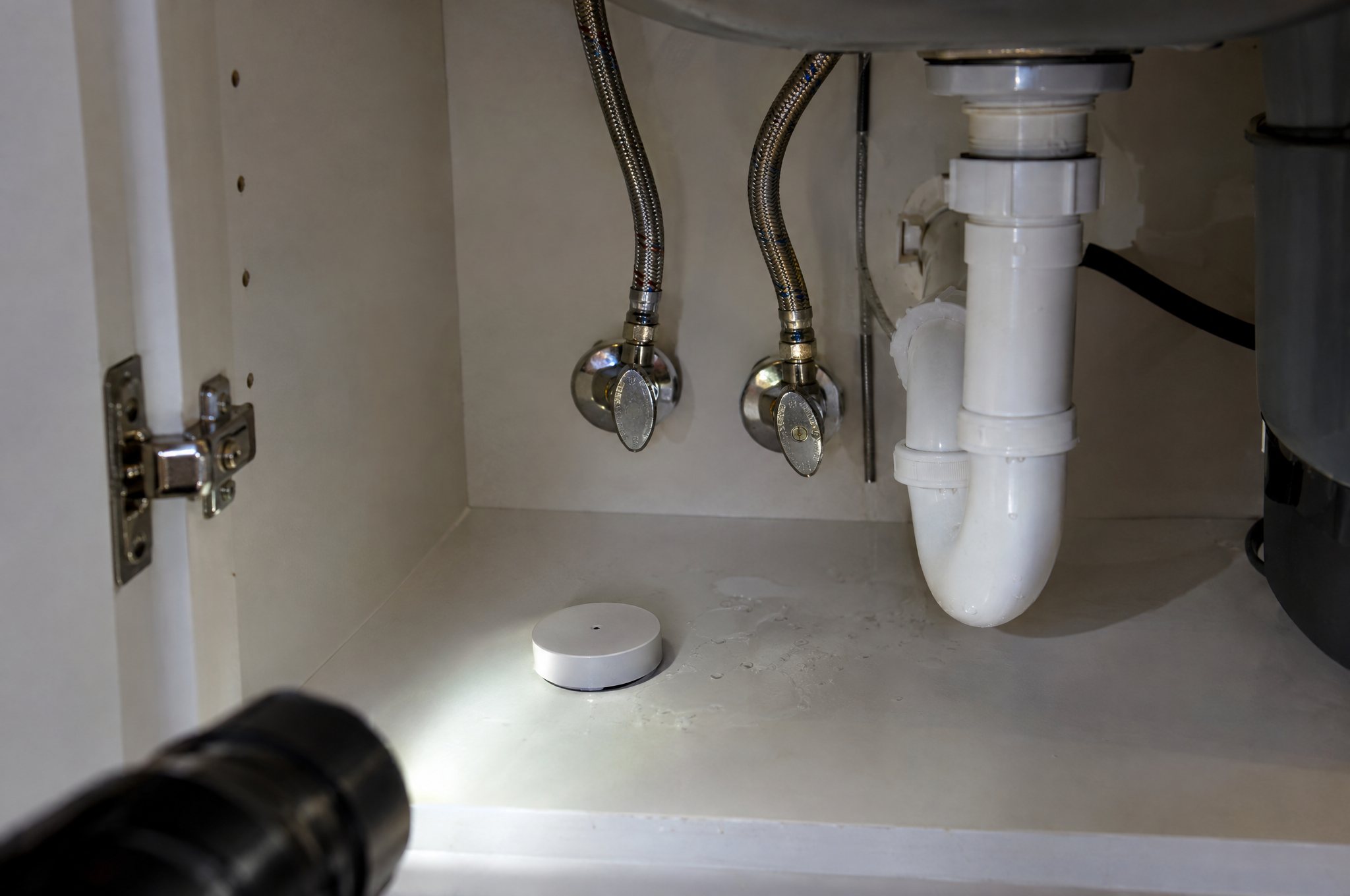 A small round white water leak sensor on the floor of a kitchen sink cabinet beside the supply lines, with a few drops of water nearby, lit by a flashlight
