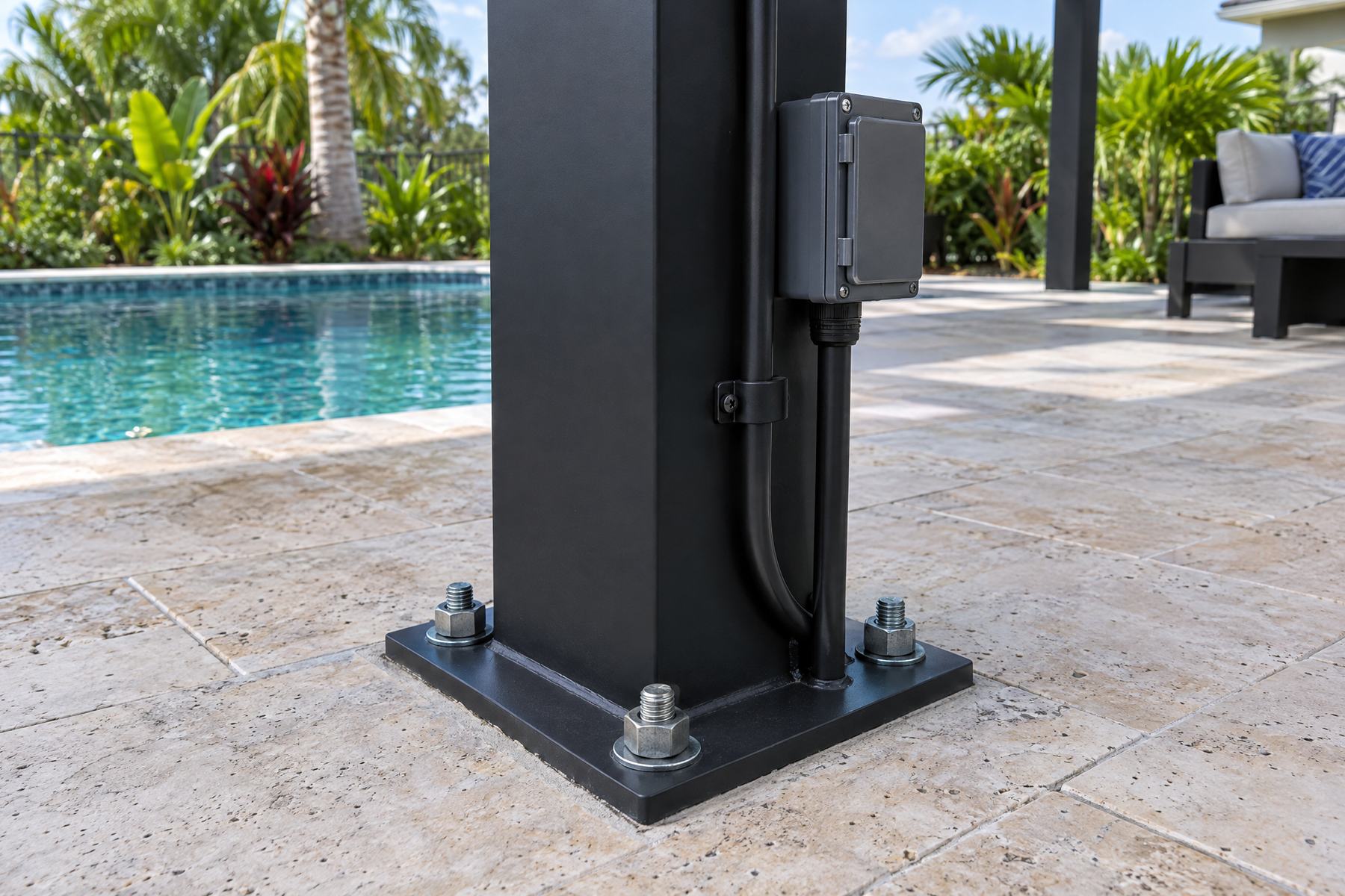 The base of a black aluminum solar pergola post bolted to a concrete footing with heavy anchor bolts, with electrical conduit running into a disconnect